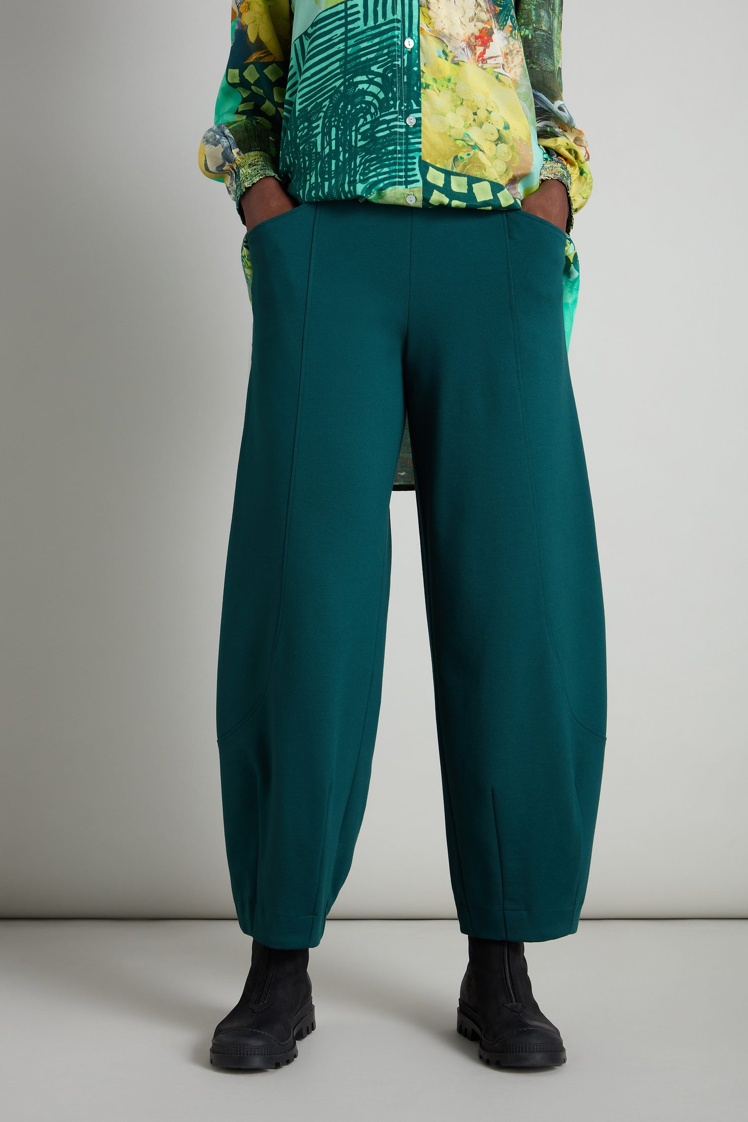 A person is dressed in a vibrant shirt and a pair of Jersey Bubble Trousers in teal, with their hands casually in the pockets, complemented by sleek black boots. The ensemble showcases a Danish-inspired design that perfectly blends style and comfort.