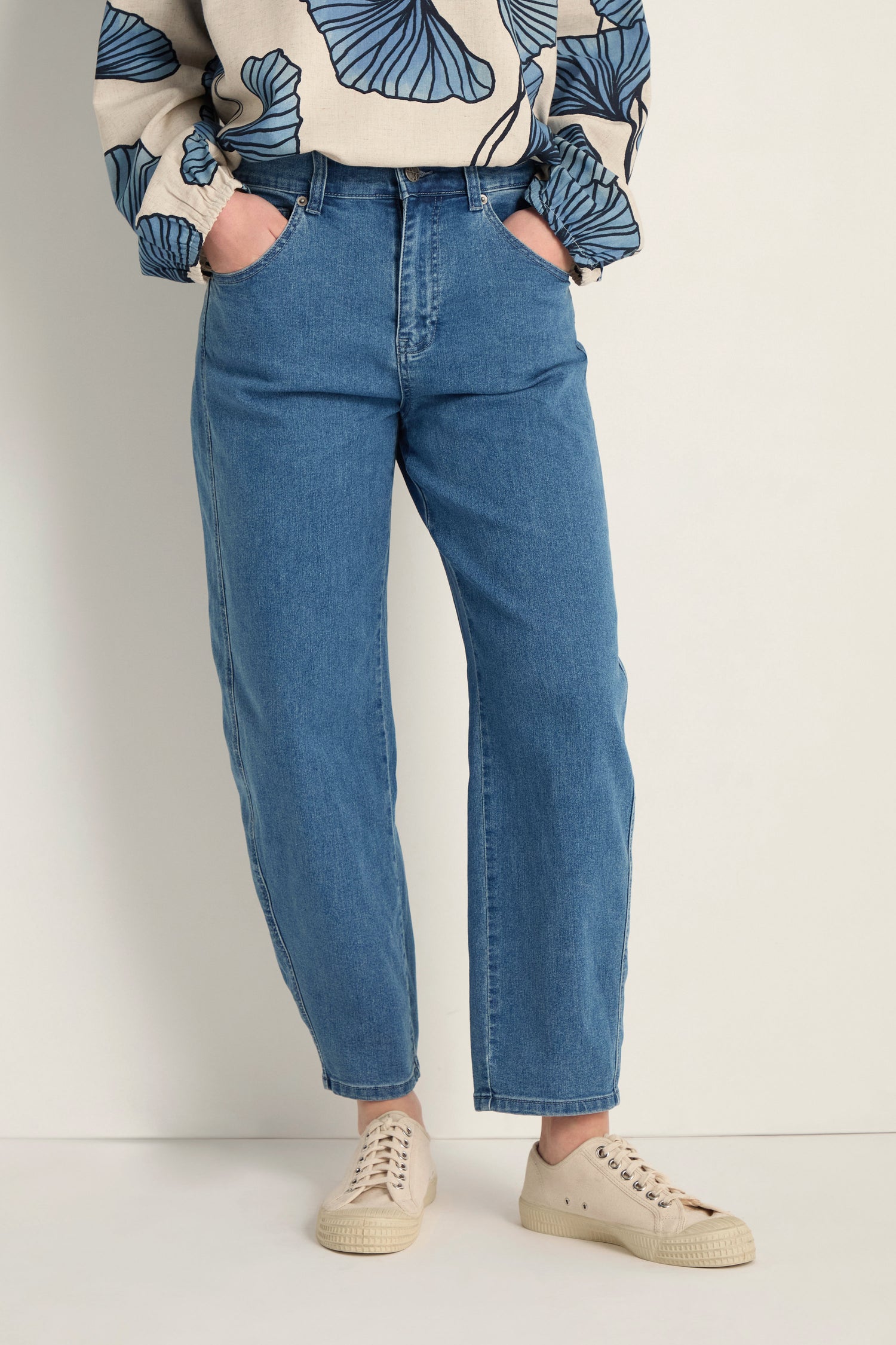 A person wearing Stretch Denim Jeans styled high-waisted and ankle-grazing, paired with a patterned long-sleeve top and beige sneakers, stands against a plain background with hands in pockets.