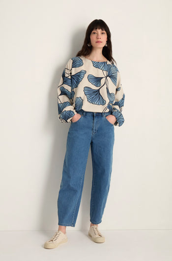 A woman stands against a plain wall, wearing a cream sweater with blue leaf patterns, Stretch Denim Jeans with a five-pocket design that graze the ankles, and beige sneakers.