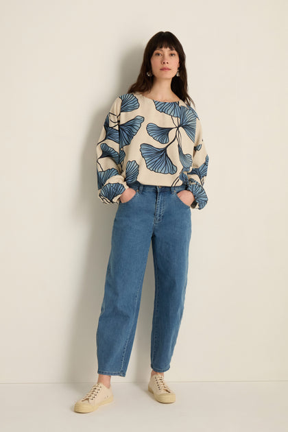 A woman stands against a plain wall, wearing a cream sweater with blue leaf patterns, Stretch Denim Jeans with a five-pocket design that graze the ankles, and beige sneakers.