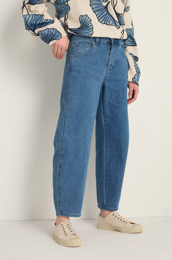 A person wears Stretch Denim Jeans in blue, featuring a five-pocket design and ankle-grazing length, paired with a beige sweater with blue leaf patterns and beige sneakers, standing against a light background.