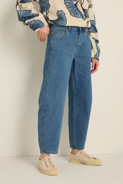 A person wears Stretch Denim Jeans in blue, featuring a five-pocket design and ankle-grazing length, paired with a beige sweater with blue leaf patterns and beige sneakers, standing against a light background.