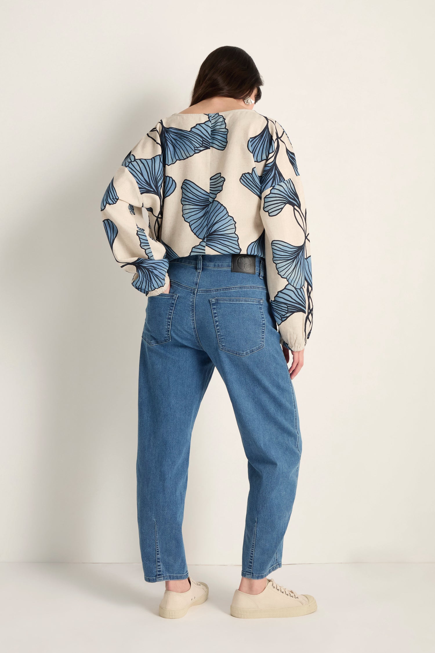 A person stands facing away from the camera, wearing a patterned long-sleeve top, beige sneakers, and Stretch Denim Jeans with a five-pocket design and ankle-grazing length against a plain, light-colored background.