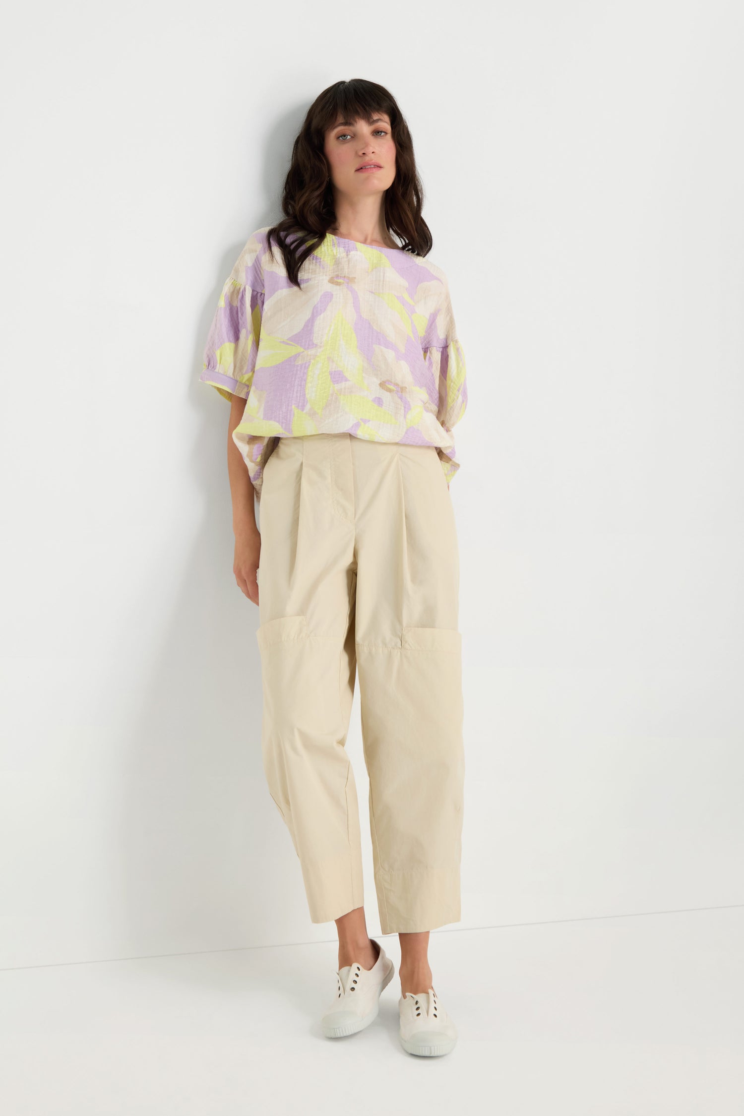 Woman stands against a white wall wearing a loose lavender and yellow patterned top, Poplin Pleated Trousers in beige high-waisted straight leg cropped style, and white sneakers.