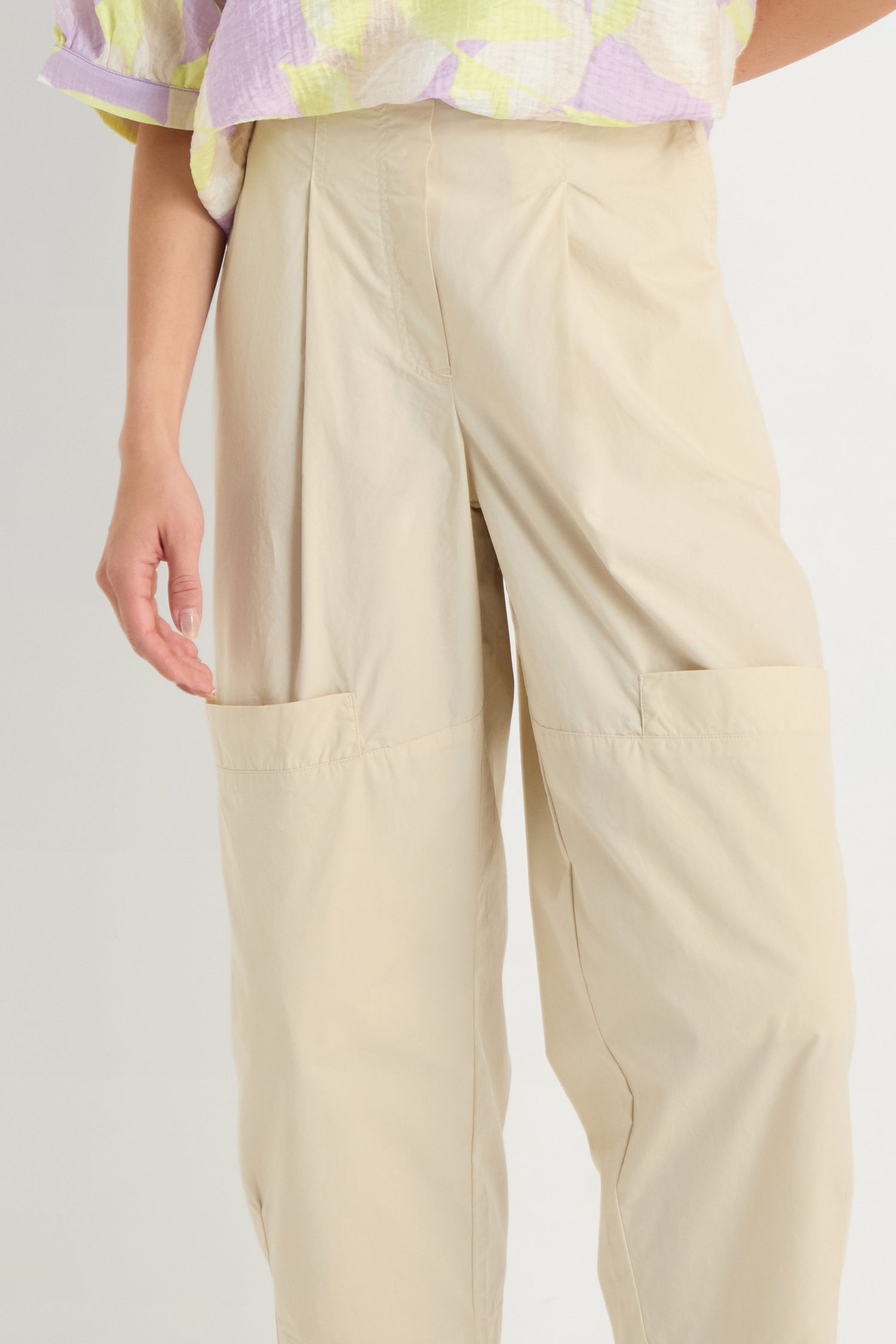 A person wearing Poplin Pleated Trousers in beige cotton with large front pockets and a pastel patterned top.