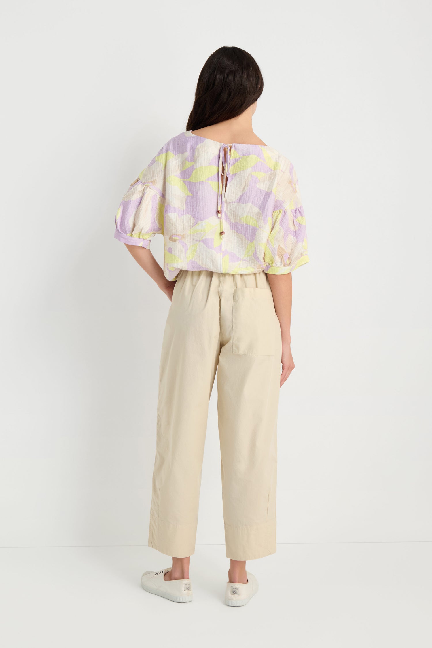 A person stands facing away, wearing pastel-patterned cotton poplin blouse, Poplin Pleated Trousers in beige, and white sneakers against a plain white background.