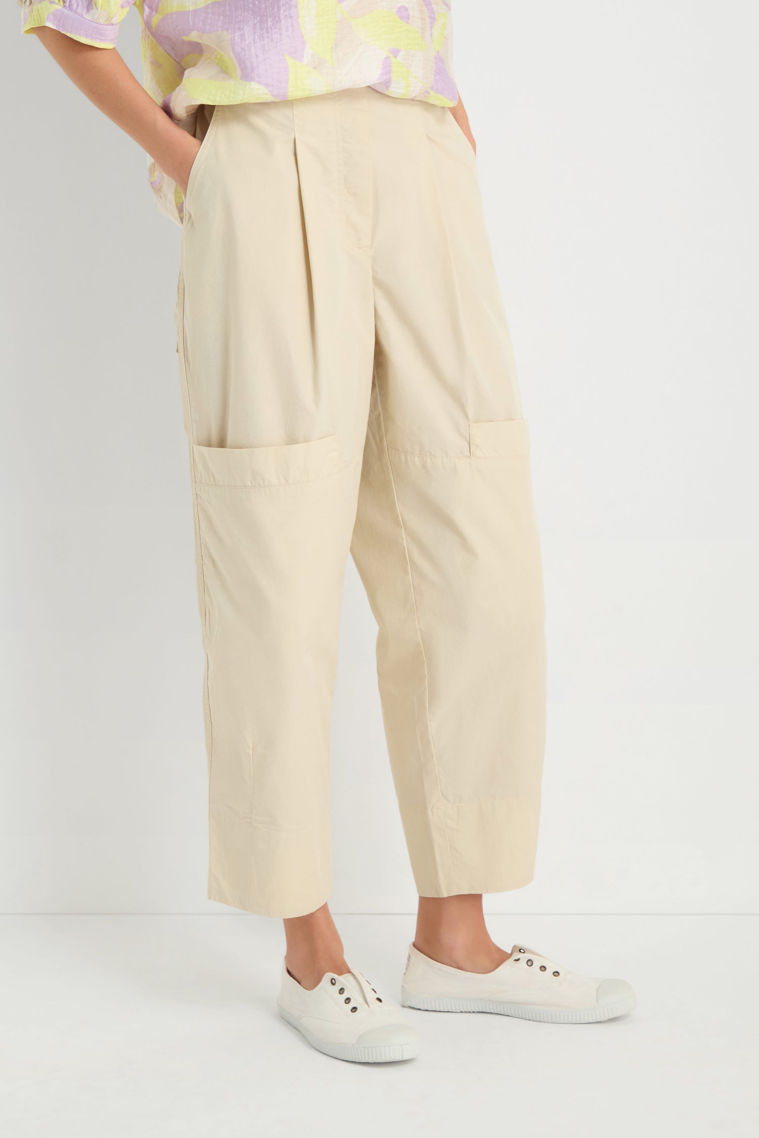 Someone stands against a white background wearing Poplin Pleated Trousers in beige with front pockets, paired with a pastel patterned shirt and white slip-on shoes.