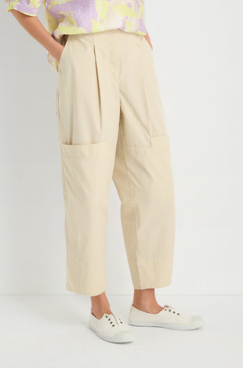 Someone stands against a white background wearing Poplin Pleated Trousers in beige with front pockets, paired with a pastel patterned shirt and white slip-on shoes.