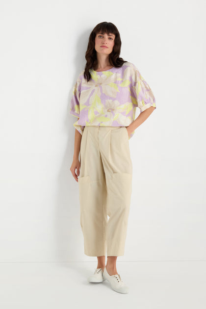 A woman stands against a white wall wearing a pastel floral blouse, Poplin Pleated Trousers with a wide straight leg in beige, and white sneakers, with one hand in her pocket.