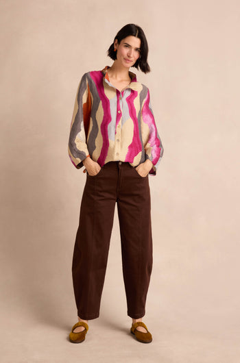 A person stands with their hands in their pockets, wearing a colorful patterned shirt, Stretch Cotton Trousers in a relaxed brown fit, and mustard yellow shoes against a plain beige background.