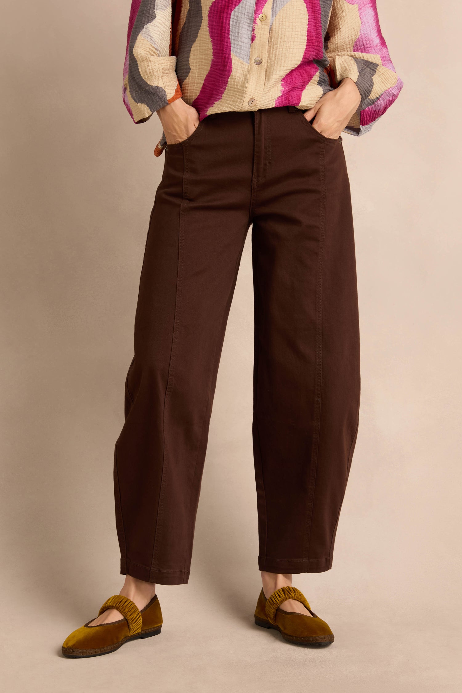 A person wearing the Stretch Cotton Trousers in brown, styled with a colorful long-sleeve shirt and mustard yellow shoes, stands with hands in pockets against a neutral background.