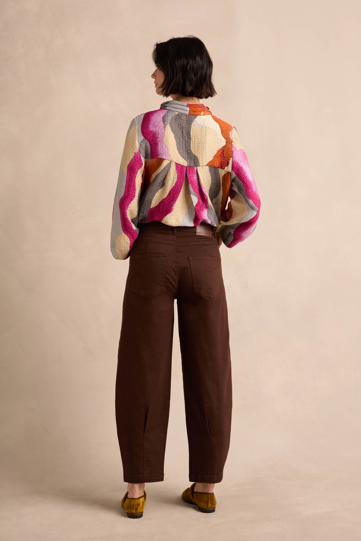 A person stands facing away, wearing relaxed fit brown Stretch Cotton Trousers, along with a colorful patterned long-sleeve shirt and mustard yellow shoes, set against a plain beige background.