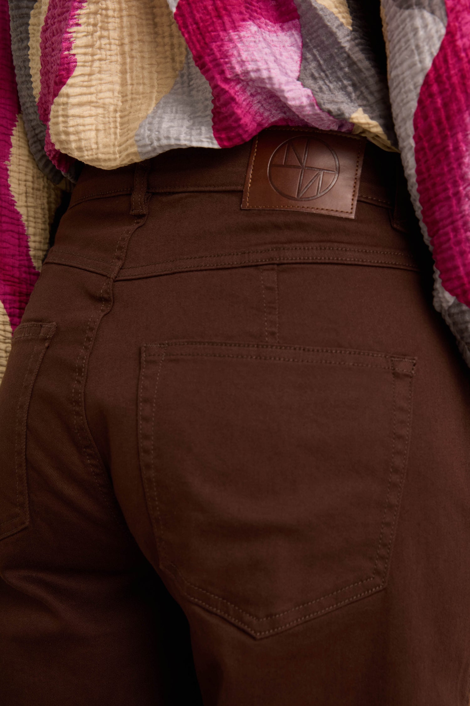 Close-up of a person wearing Relaxed Fit Stretch Cotton Trousers in brown with a leather logo patch on the waistband, paired with a textured, multicolored top.