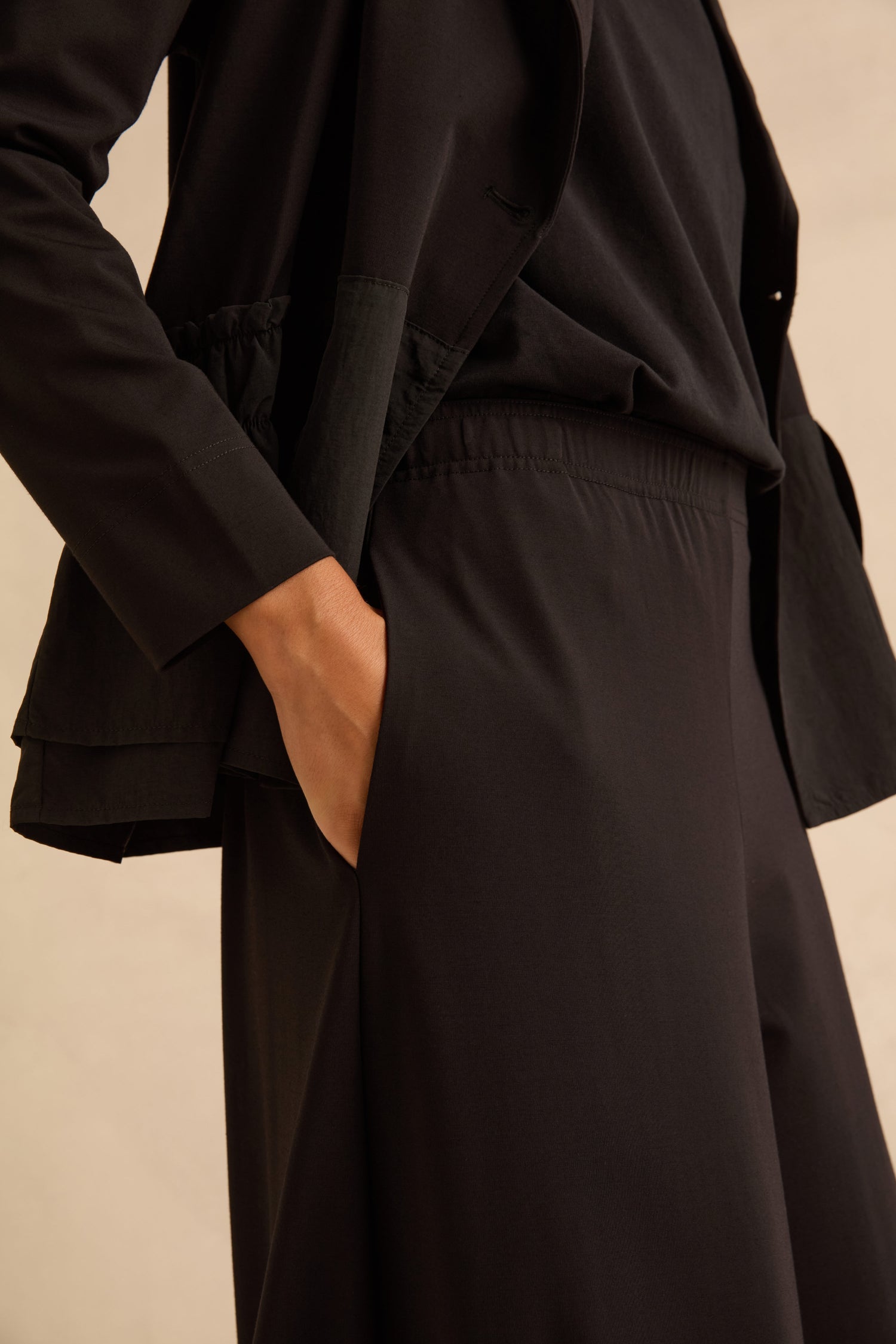 Person wearing Black Pleated Hem Wide Jersey Trousers with a cropped leg and elastic waistband with a hand in the pocket, paired with a matching black jacket.