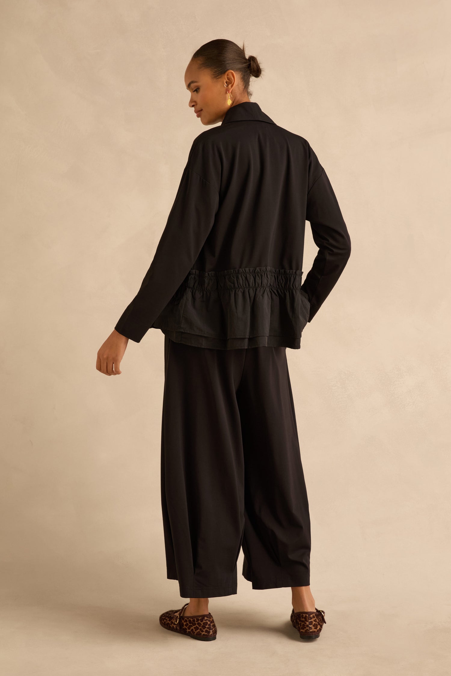 A model wears a black collared jacket with a pleated peplum hem and Black Pleated Hem Wide Jersey Trousers, styled with leopard-print loafers.