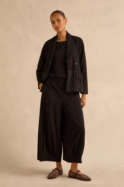 A model wears a black jacket with a flower detail, a black top, and Black Pleated Hem Wide Jersey Trousers, paired with leopard-print flats.