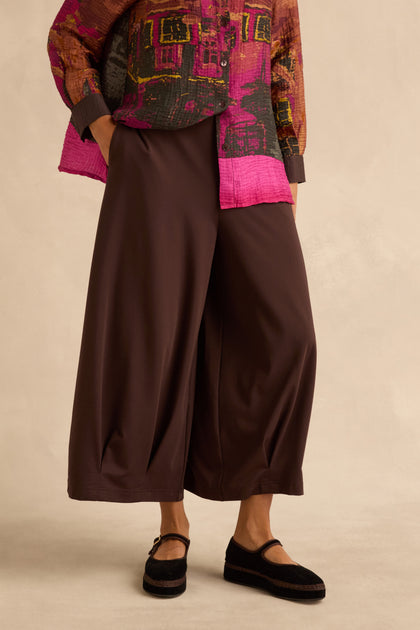 A person wears Chocolate Pleated Hem Wide Jersey Trousers with a patterned button-down shirt and black mary jane shoes.