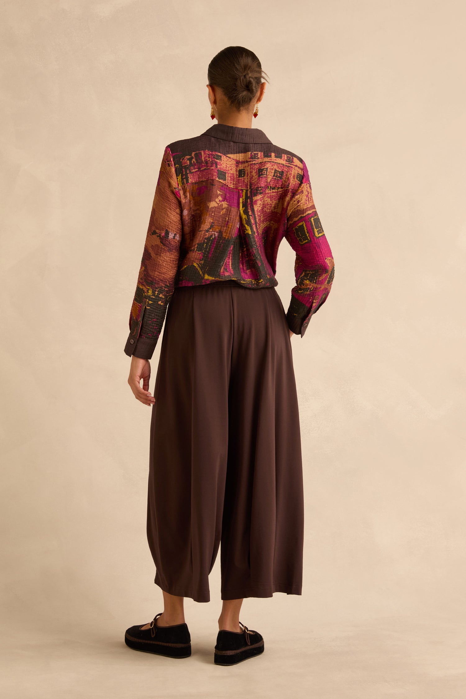 A person viewed from behind, wearing a long-sleeved button-down shirt with a multicolored abstract print and Chocolate Pleated Hem Wide Jersey Trousers.