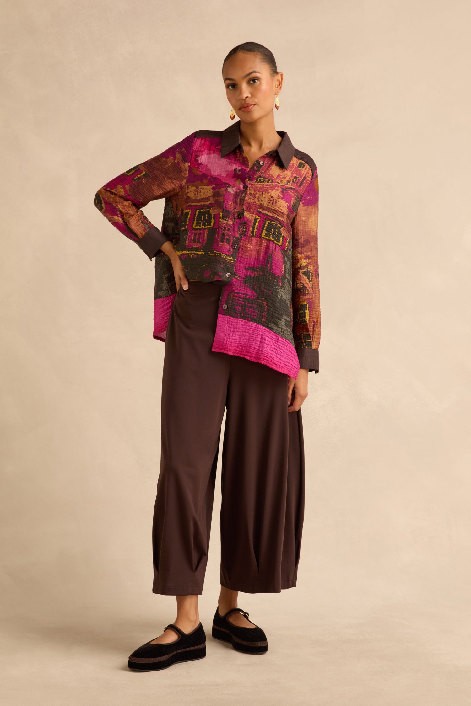 A person wearing a long-sleeved button-down shirt with a vibrant abstract print and Chocolate Pleated Hem Wide Jersey Trousers.