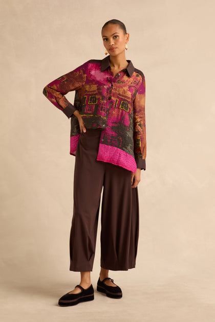 A person wearing a long-sleeved button-down shirt with a vibrant abstract print and Chocolate Pleated Hem Wide Jersey Trousers.