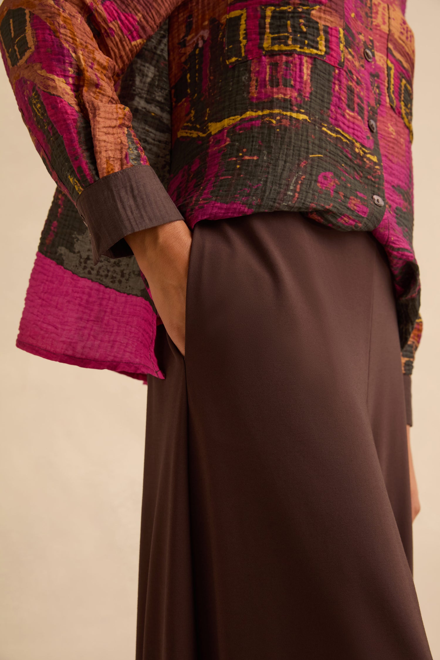 A person wears a long-sleeved shirt with a purple, black, and yellow block-print pattern and Chocolate Pleated Hem Wide Jersey Trousers with a hand in the pocket.