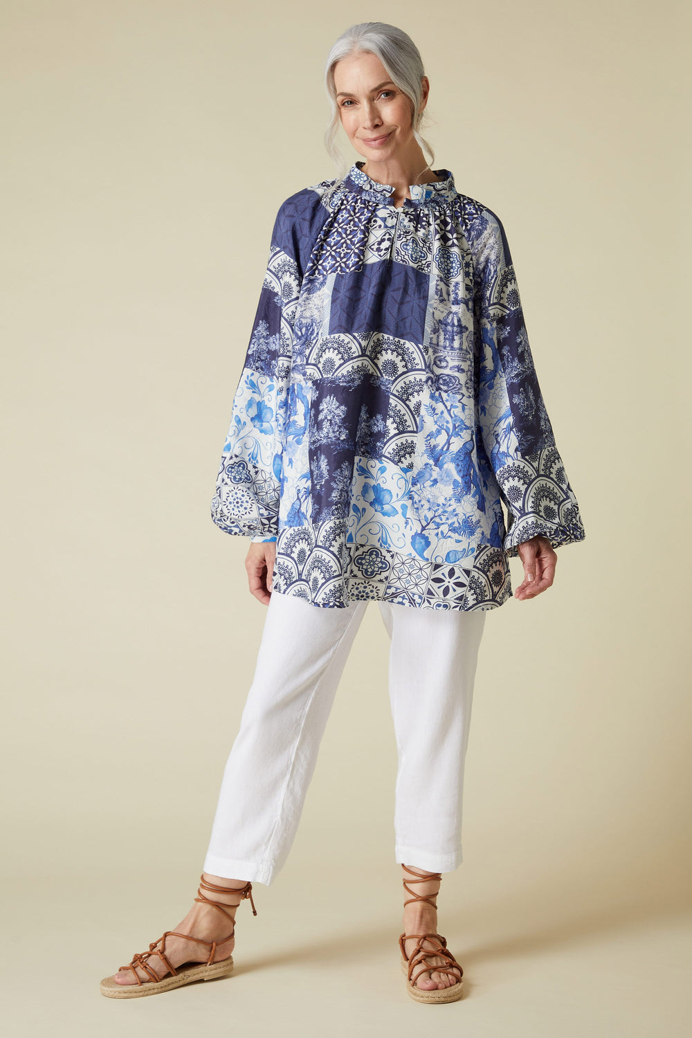 A woman posing in a Long Sleeve Porcelain Print Top featuring bell sleeves, styled with white pants and brown sandals.