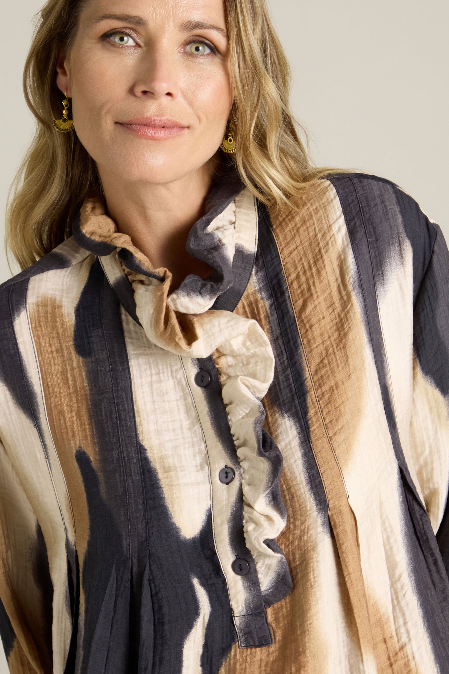 A person with long hair is wearing the Crinkle Viscose Print Blouse, which features a black, brown, and cream pattern along with ruffles and buttons. They are looking slightly off-camera and smiling subtly.