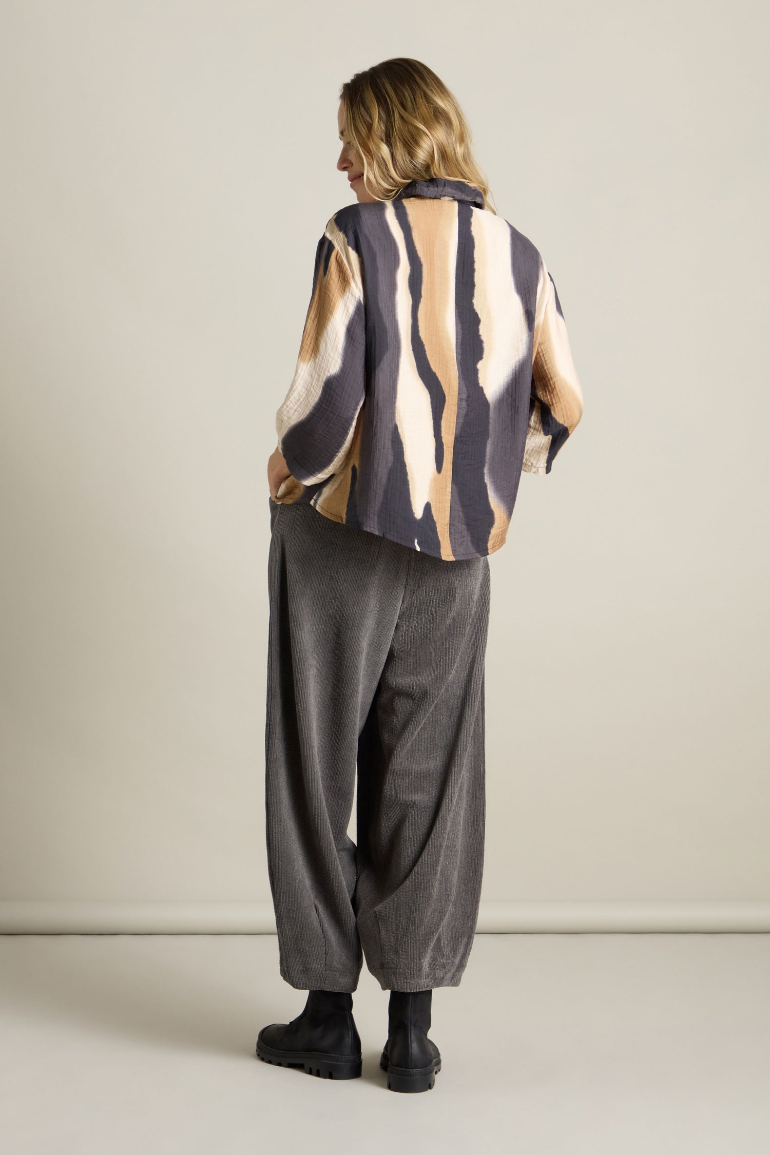 Person stands with their back to the camera, wearing a Crinkle Viscose Print Blouse featuring a loose watercolor pattern, wide gray pants, and black boots.