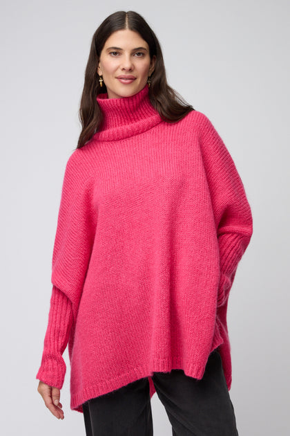 A woman wears the Pink Wool Mix Poncho, a loose-fitting modern knitwear in bright pink with long sleeves, paired with dark pants, standing against a neutral background.