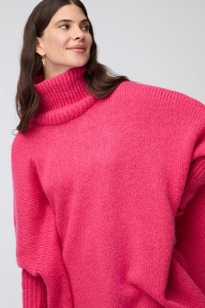 A woman with long brown hair wears a Pink Wool Mix Poncho and gold earrings, standing against a plain light gray background.