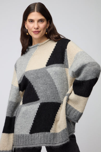 A woman models the Grey Colour Block Knit, a loose sweater in grey, black, and beige tones, embodying Scandinavian minimalism against a light background.