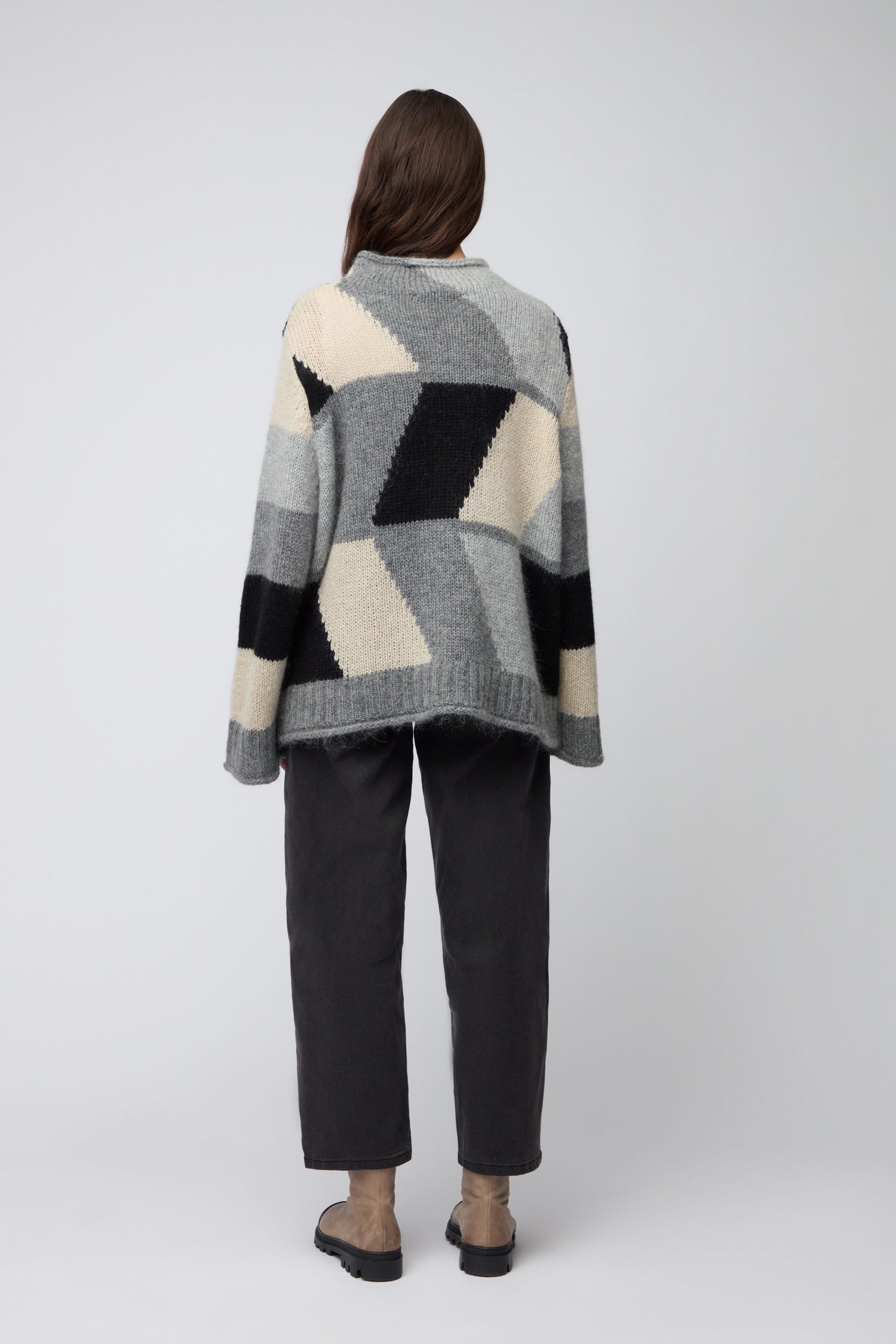 A person stands facing away in the Grey Colour Block Knit—shades of grey, black, and cream—paired with dark pants and brown shoes against a plain background, channeling effortless Scandinavian minimalism.