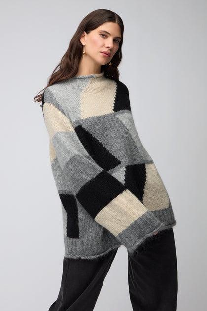 A woman with long brown hair wears the Grey Colour Block Knit in gray, black, and beige with dark pants, standing against a plain light background for a Scandinavian minimalist look.