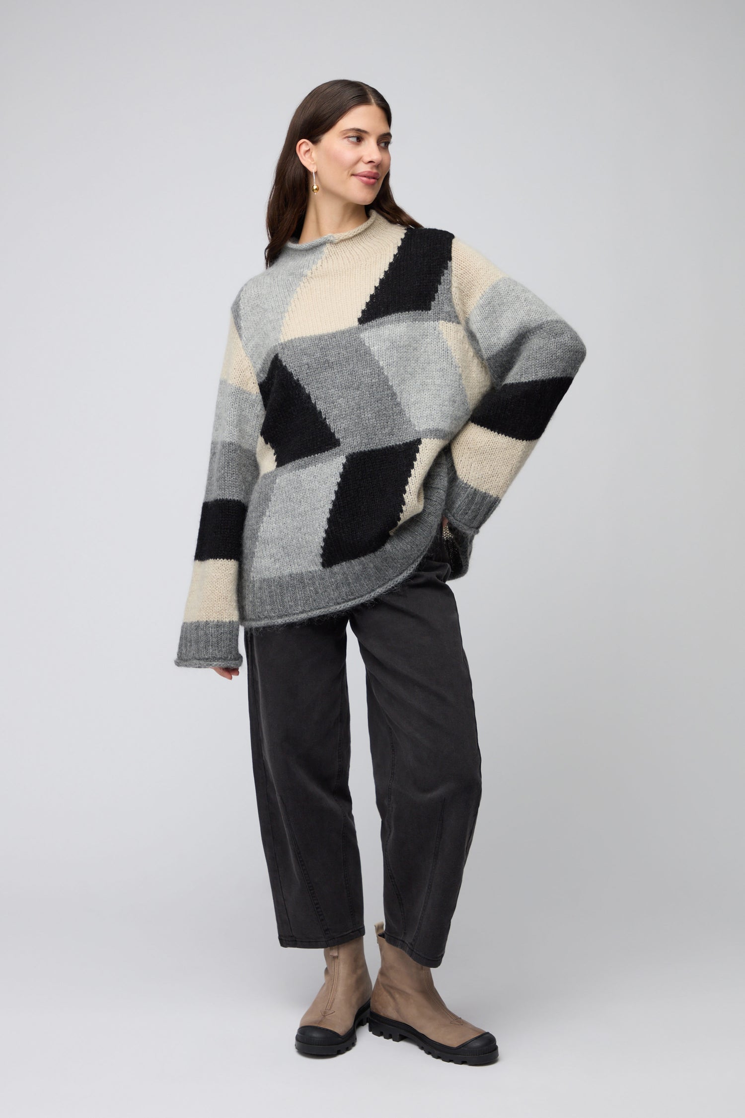 A woman stands against a plain background wearing the Grey Colour Block Knit, loose dark pants, hoop earrings, and beige shoes—an effortless nod to Scandinavian minimalism.