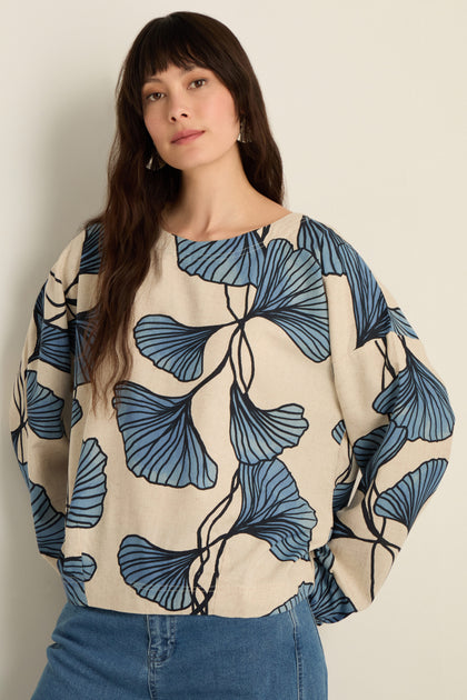 A woman stands against a plain wall, wearing the Ginkgo Viscose Linen Boxy Top with large blue leaf patterns and a blue denim skirt.