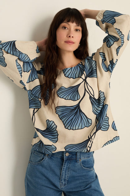 A woman stands against a white wall wearing a cream Ginkgo Viscose Linen Boxy Top with blue leaf patterns and blue jeans, her arms raised and hands resting on her head.