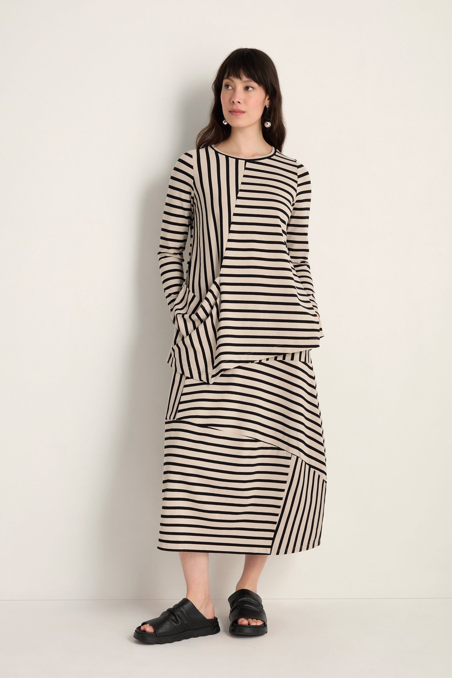 A woman stands against a plain wall wearing the Asymmetric Stripe Jersey Tunic in beige and black with a matching skirt, both crafted from soft viscose jersey, paired with black sandals.