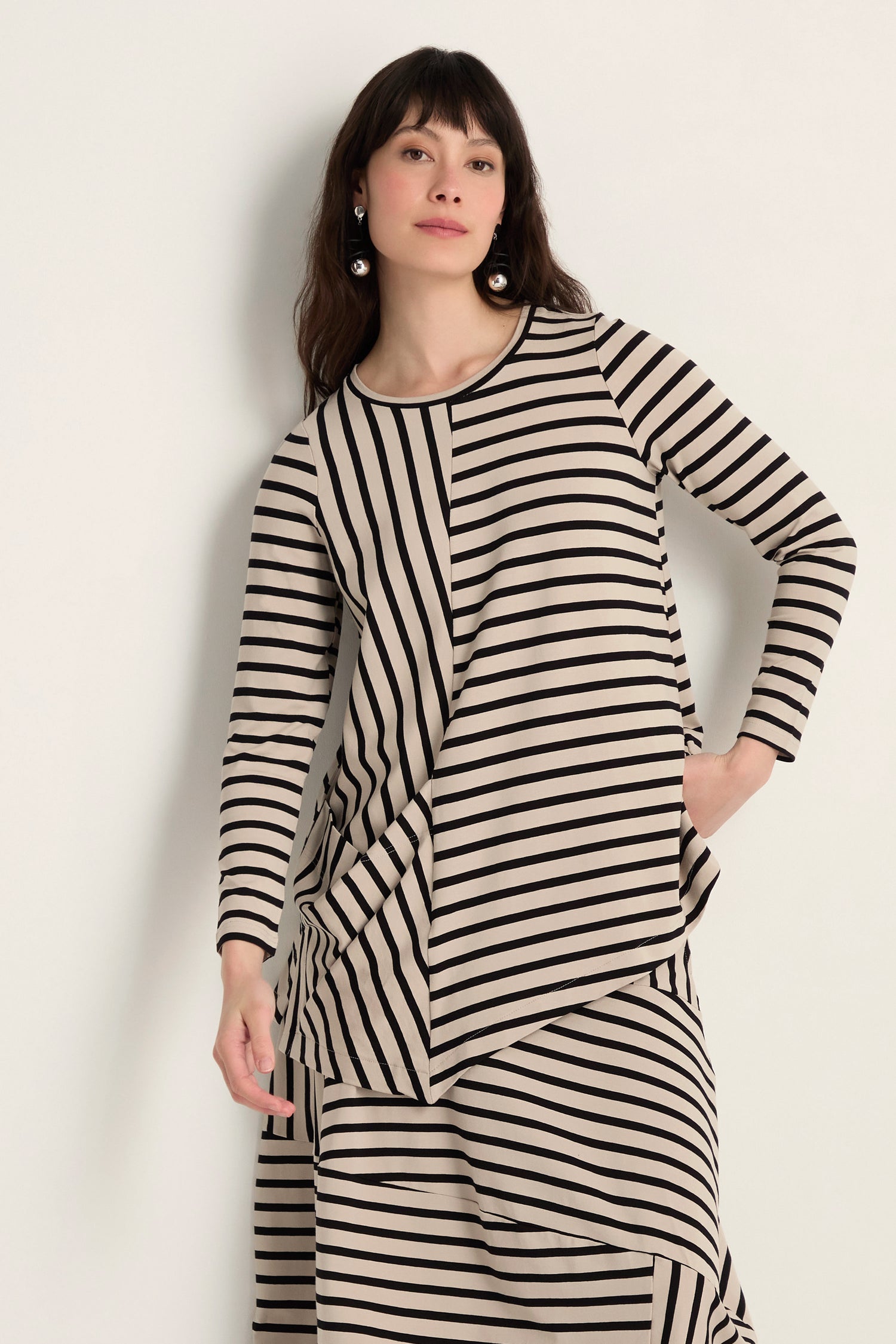 A woman models the Asymmetric Stripe Jersey Tunic—a long-sleeved, beige and black piece with an asymmetrical hem—paired with a matching skirt, set against a plain light background.
