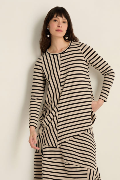 A woman models the Asymmetric Stripe Jersey Tunic—a long-sleeved, beige and black piece with an asymmetrical hem—paired with a matching skirt, set against a plain light background.