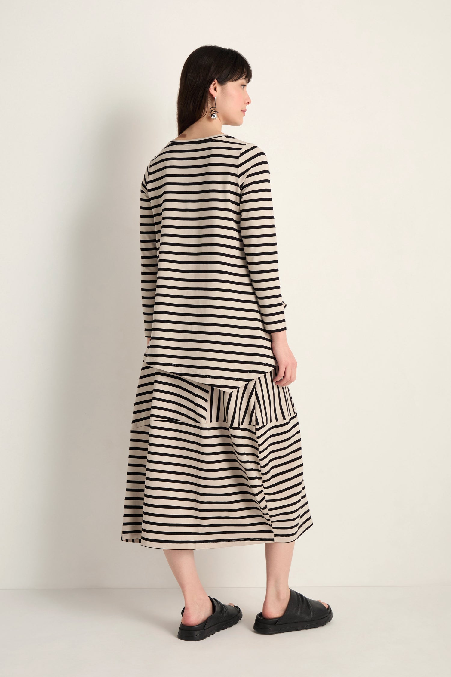 Woman stands against a plain background, facing away, wearing the Asymmetric Stripe Jersey Tunic in black and beige stripes with matching skirt and black sandals.