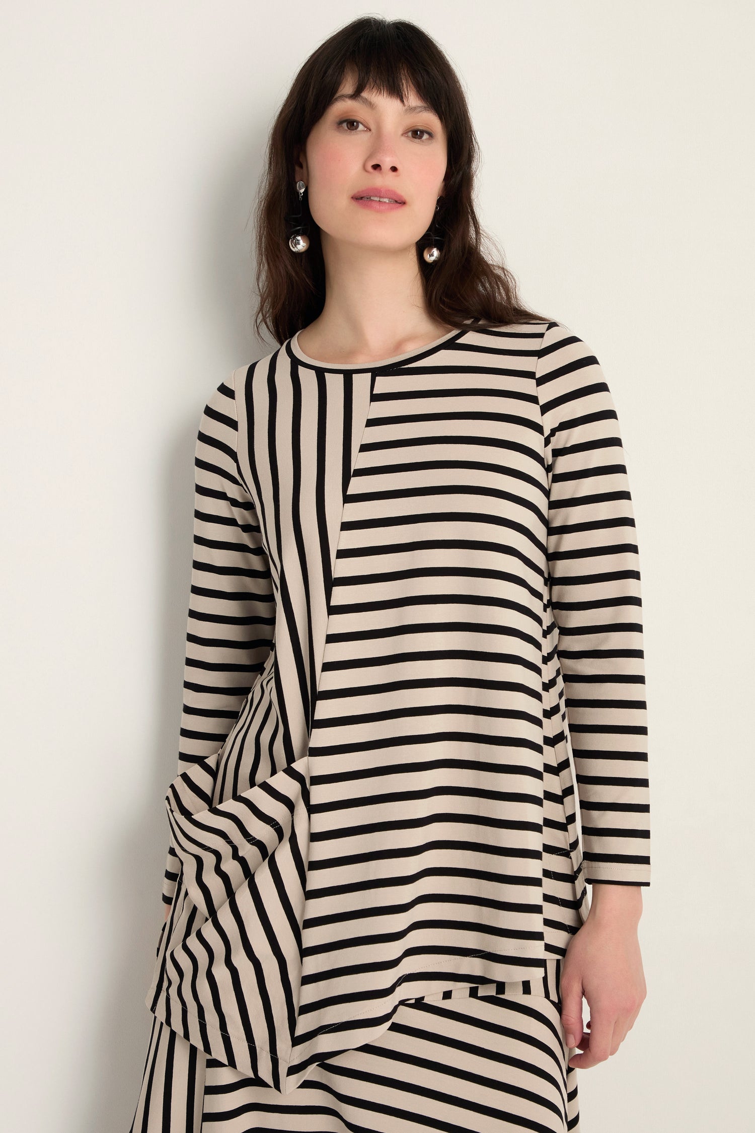 Woman in an Asymmetric Stripe Jersey Tunic with long sleeves and a draped hem, made from soft viscose jersey, poses against a plain light-colored wall.