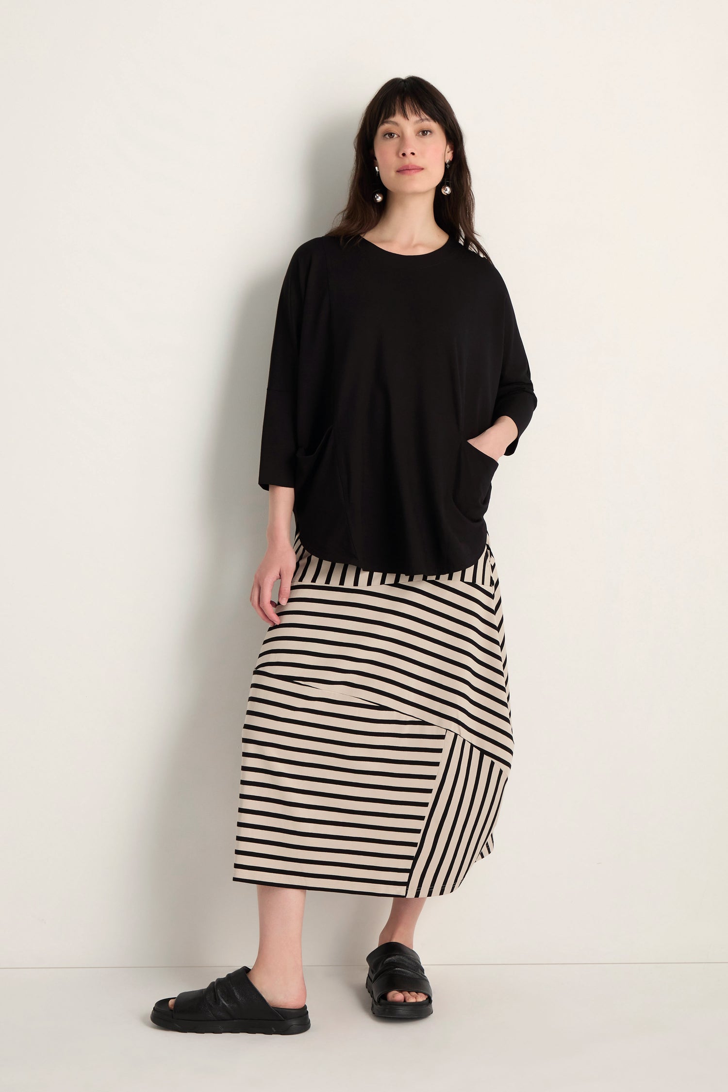 A woman stands against a light wall, wearing the Batwing Jersey Top in black with a relaxed drape, paired with a beige and black striped viscose jersey skirt and black sandals. One hand is tucked in her pocket.