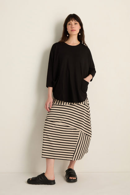 A woman stands against a light wall, wearing the Batwing Jersey Top in black with a relaxed drape, paired with a beige and black striped viscose jersey skirt and black sandals. One hand is tucked in her pocket.