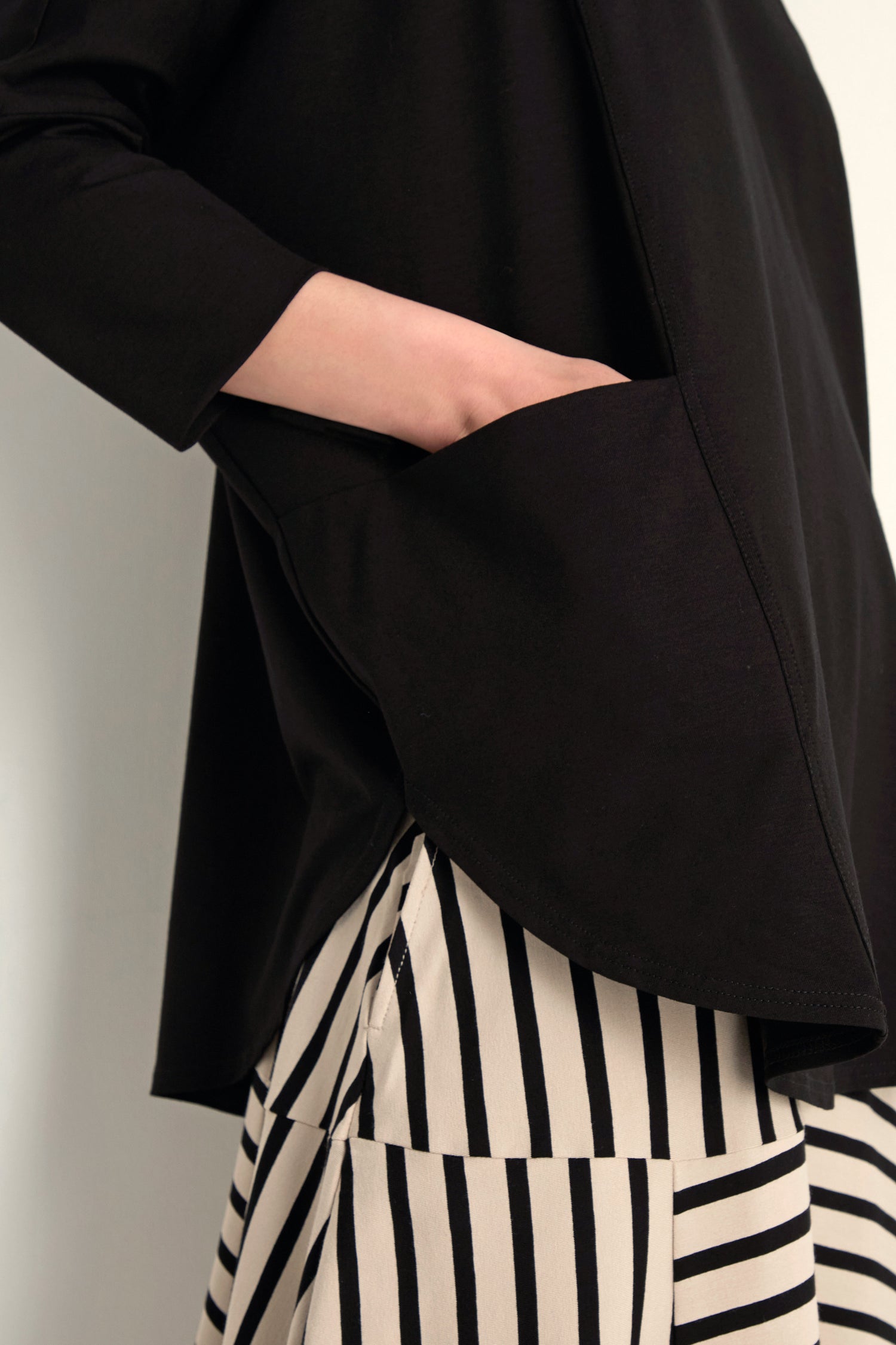 A person wearing the Batwing Jersey Top in black with one hand in their pocket, paired with cream pants featuring black vertical stripes.