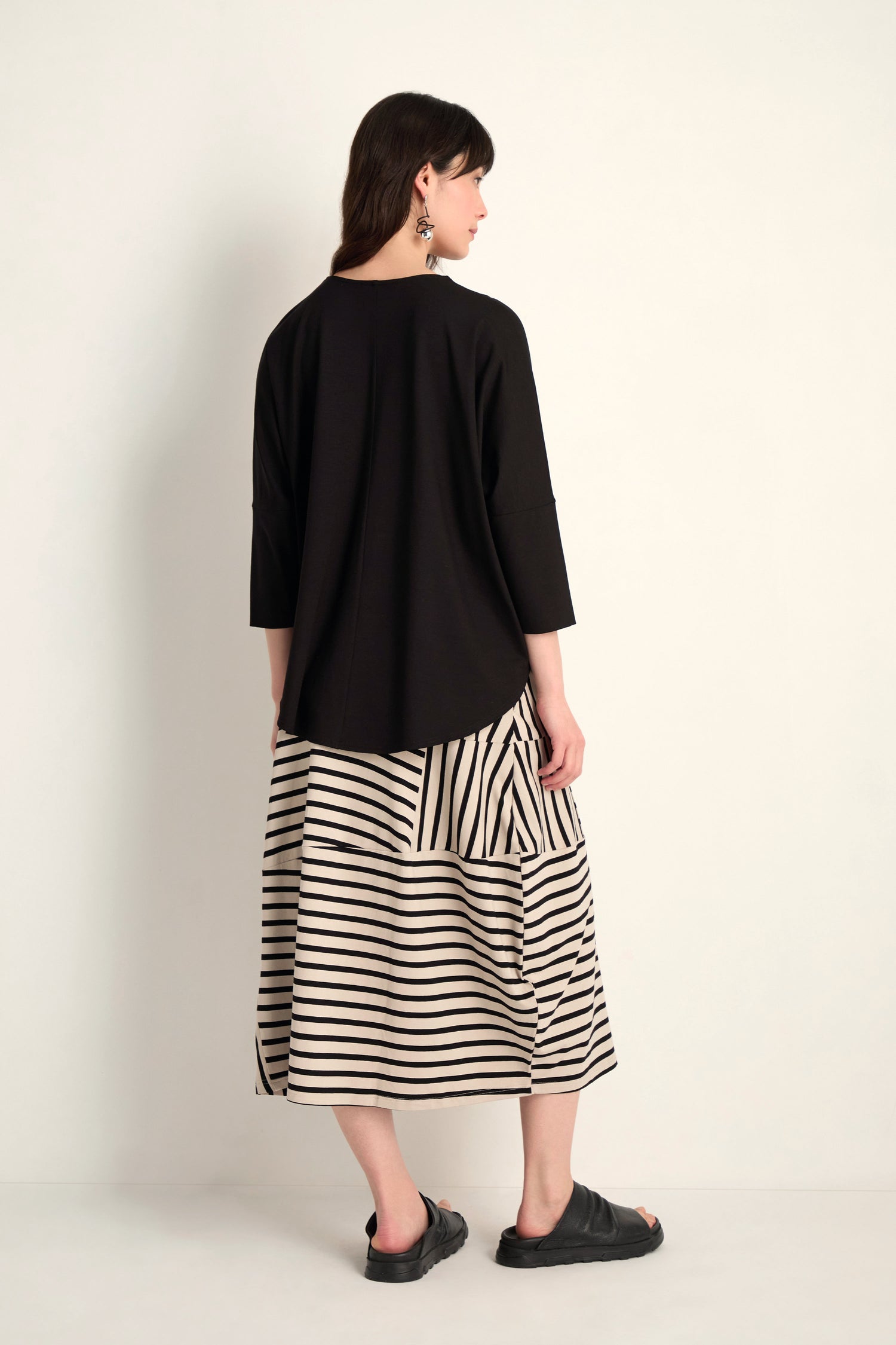 A woman stands against a plain background, facing away, wearing the Batwing Jersey Top in black with a lightweight drape, paired with a beige and black striped midi skirt and black sandals.