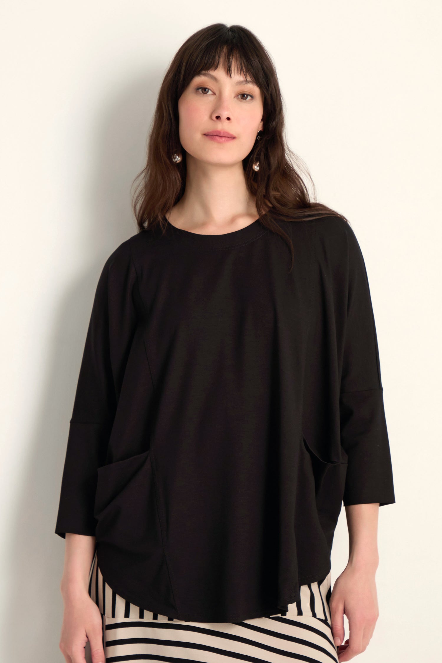 A woman with long brown hair wears the Batwing Jersey Top, featuring front pockets and a lightweight viscose jersey drape, paired with a black-and-white striped skirt against a plain light background.
