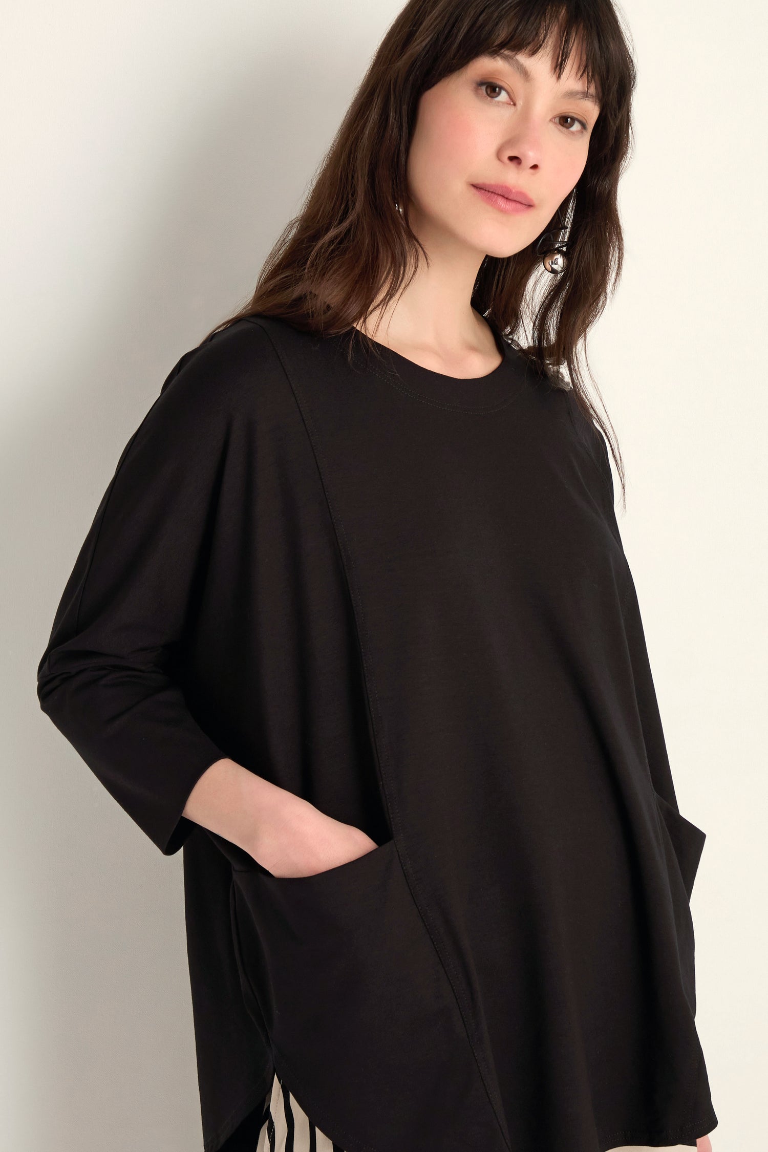 A woman wears the Batwing Jersey Top, featuring large front pockets and a loose fit in lightweight viscose jersey, as she stands against a light wall and looks at the camera.