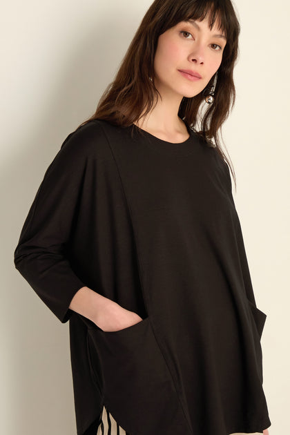 A woman wears the Batwing Jersey Top, featuring large front pockets and a loose fit in lightweight viscose jersey, as she stands against a light wall and looks at the camera.