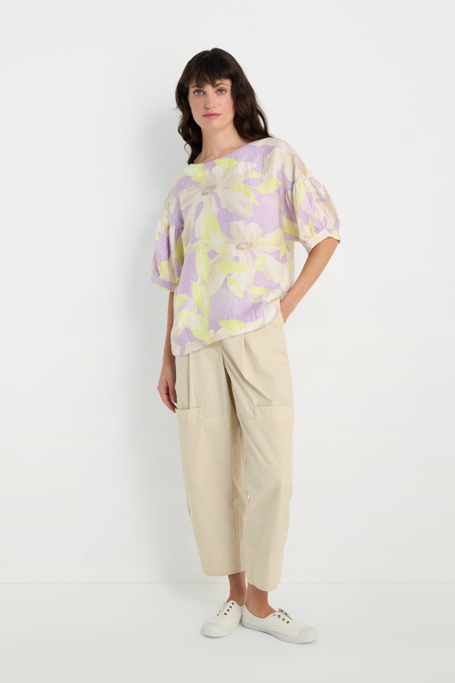 A woman stands against a white background wearing the Puff Sleeve Print Top featuring pastel florals and elbow-length puff sleeves, paired with beige wide-leg pants with pockets and white slip-on shoes.