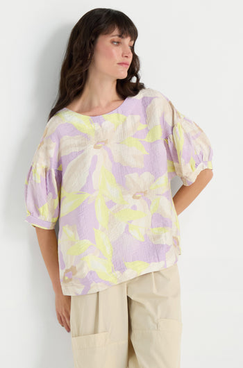 A woman in a Puff Sleeve Print Top, featuring a relaxed fit, pastel floral design, soft viscose blend, elbow-length puff sleeves, and beige pants stands against a plain white background.